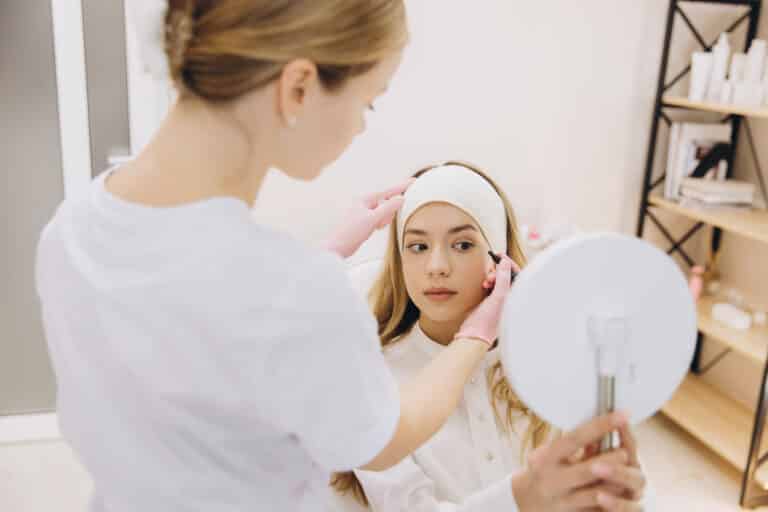 A beautician in white with pink gloves applies makeup to a young woman wearing a white headband, who looks into a mirror.