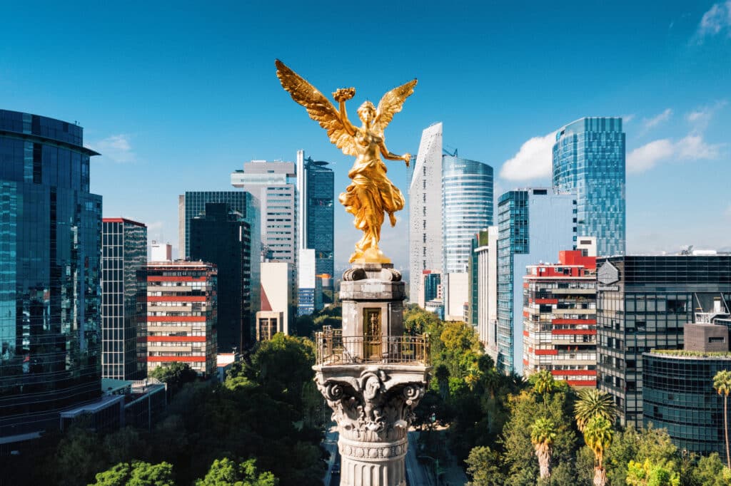 Golden statue of the winged Victoria atop a tall ornate column, set against a modern city skyline with glass skyscrapers and blue sky.