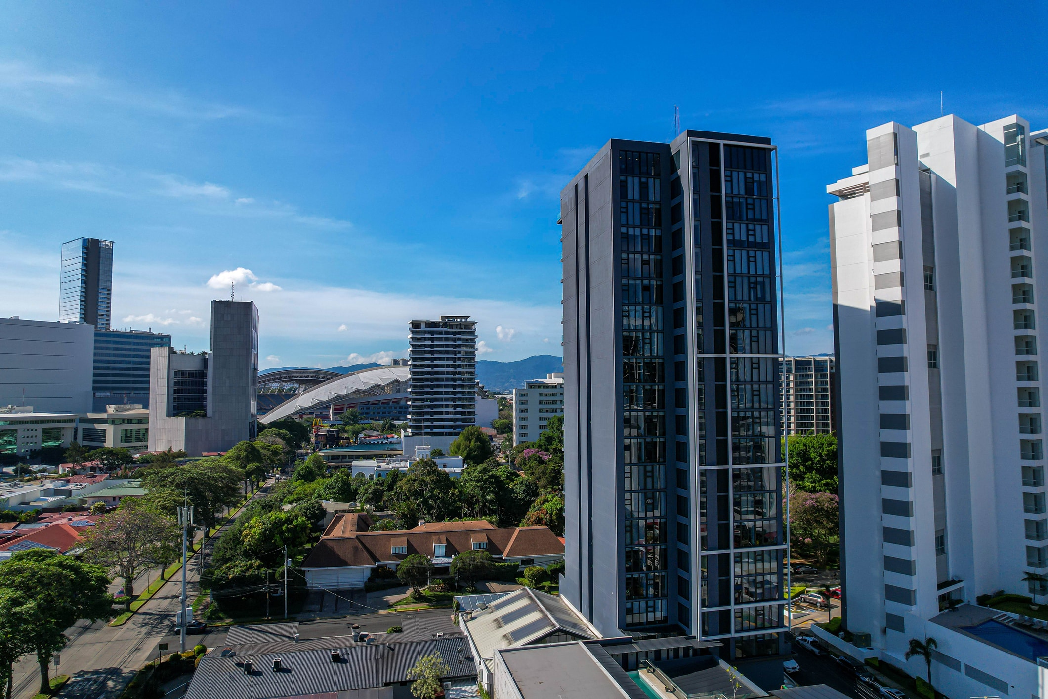 A cityscape with tall modern buildings under a clear blue sky, including a pair of dark glass towers in the foreground and a mix of lower structures and greenery below.