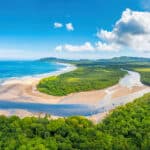 Aerial view of a tropical coastal landscape with a wide blue ocean to the left, a sandy beach curving along the shore, and a winding river through lush green forests toward distant hills under a bright blue sky with fluffy white clouds.