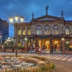 Historic ornate theater building with statues on top, lit entrance, iron fence, and a fountain in the foreground at dusk. Urban square with flag and modern buildings nearby.