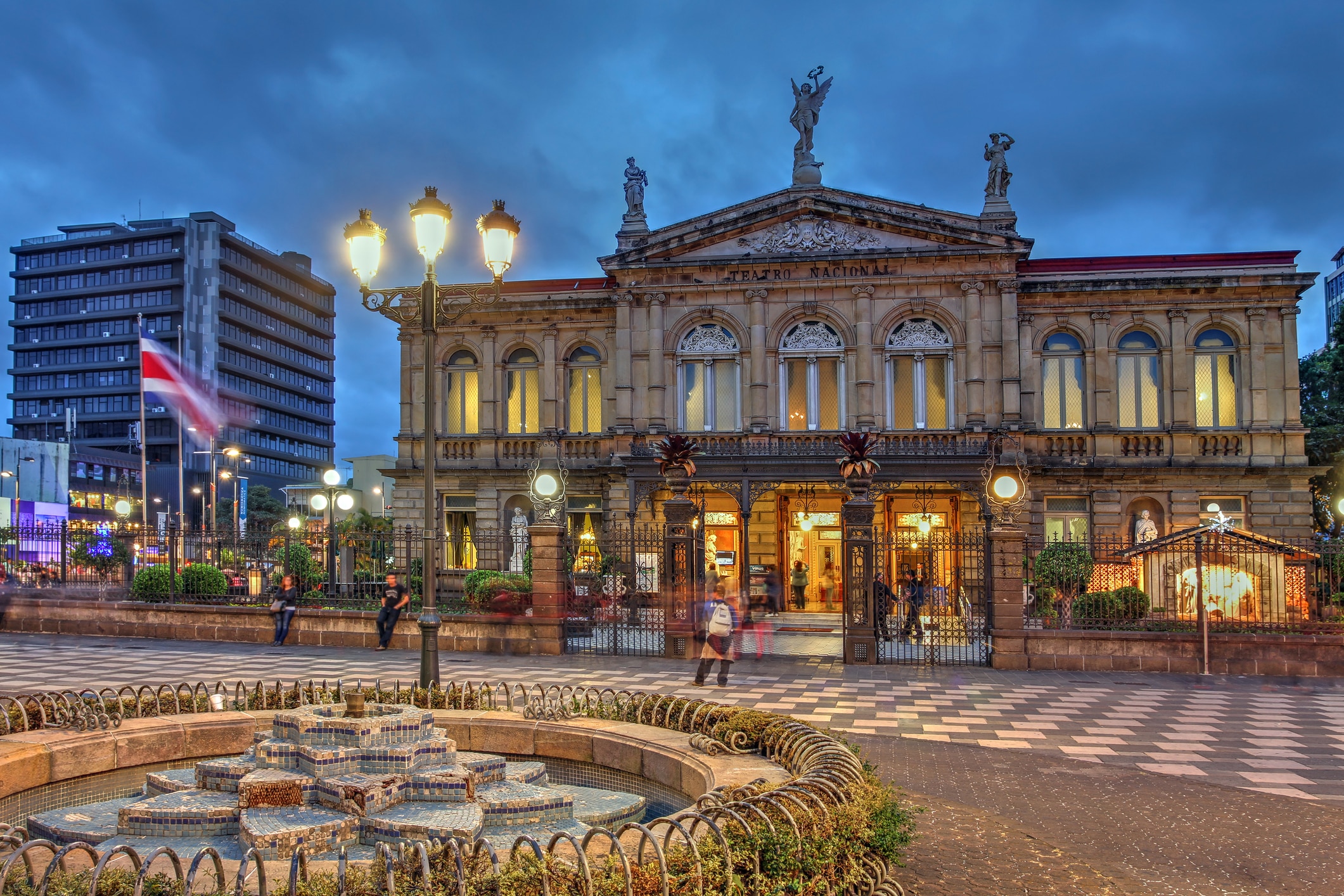 Historic ornate theater building with statues on top, lit entrance, iron fence, and a fountain in the foreground at dusk. Urban square with flag and modern buildings nearby.