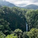 A tall waterfall plunging off a cliff into a dense tropical forest, surrounded by lush green mountains and thick foliage under a partly cloudy sky.