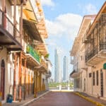 A sunlit narrow street lined with colorful colonial buildings, balconies with ironwork, and a view of modern high-rise towers in the distance under a blue sky.