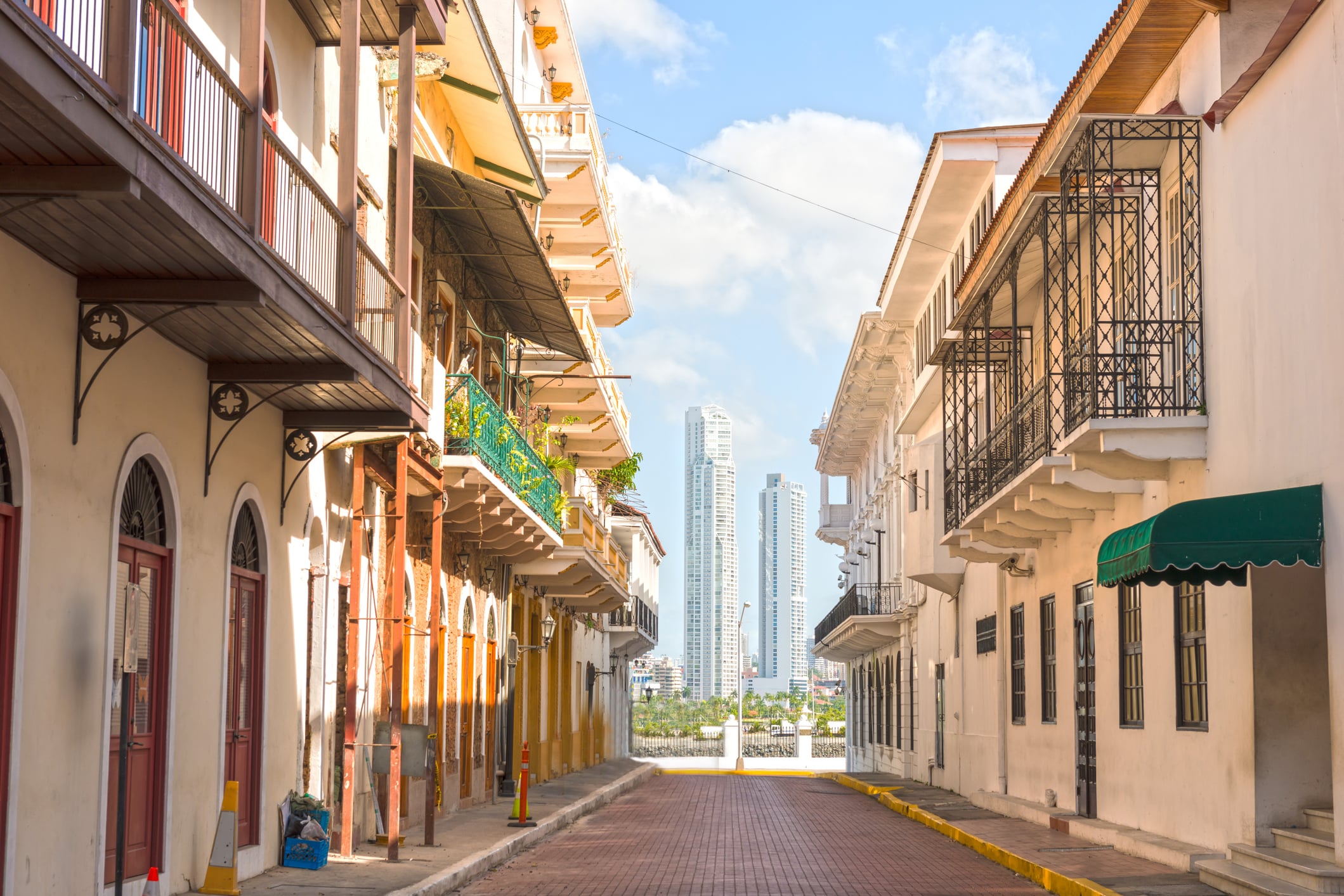 A sunlit narrow street lined with colorful colonial buildings, balconies with ironwork, and a view of modern high-rise towers in the distance under a blue sky.