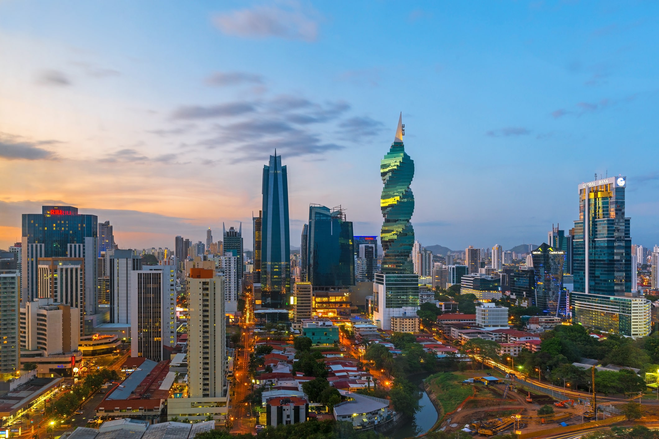 A city skyline at dusk with modern high-rise buildings, including a spiraled green tower, illuminated streets, and a river or canal cutting through a residential area in the foreground.