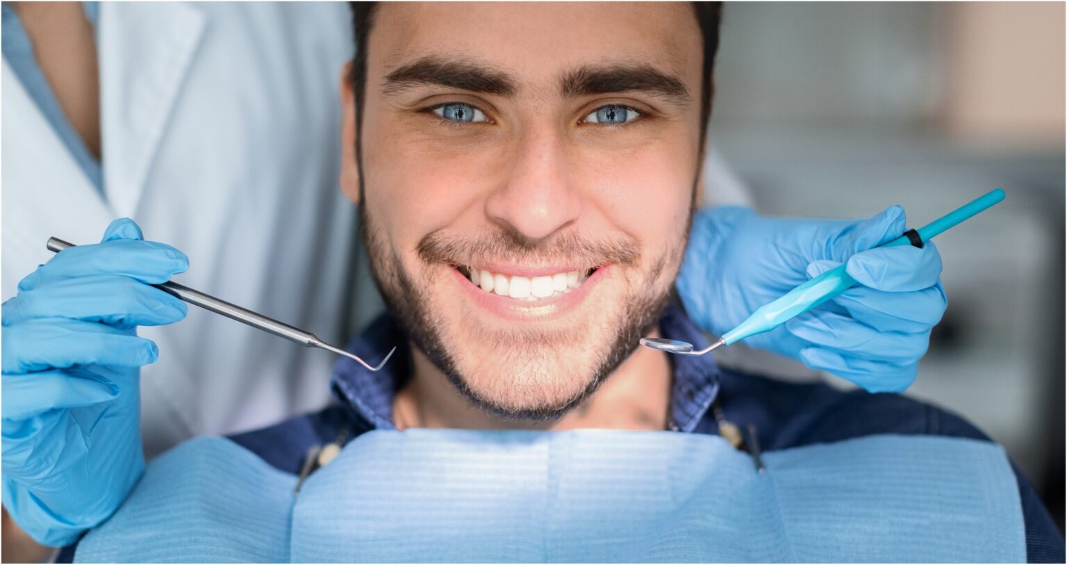 A smiling man seated in a dental chair, blue eyes and beard, being treated by a dentist with blue gloves holding dental instruments on each side.