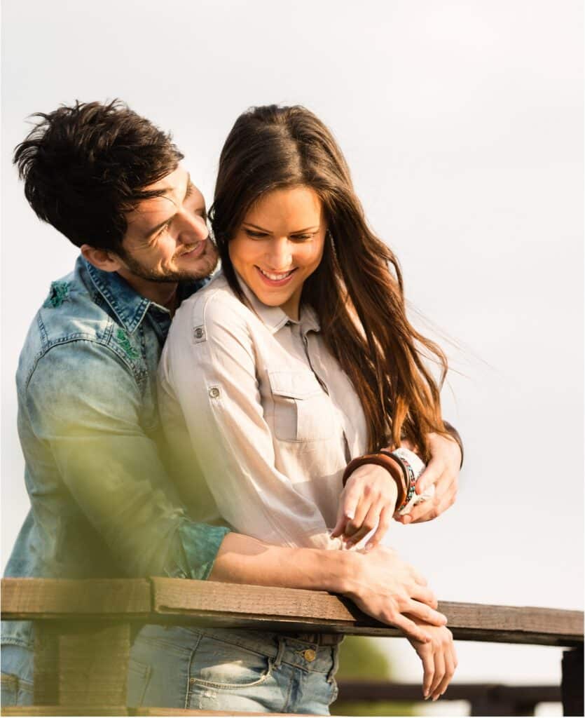 A smiling couple standing on a wooden railing, the man embracing from behind with his arms around the woman as they lean together outdoors.