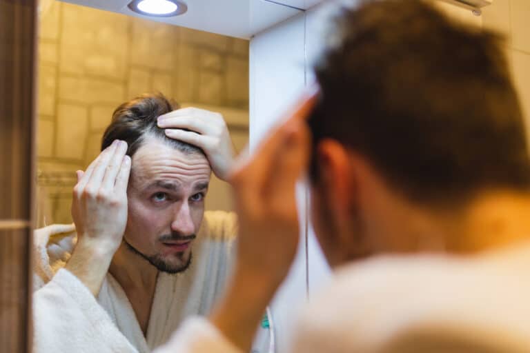 A man wearing a white bathrobe looks at himself in a bathroom mirror, running fingers through his hair.