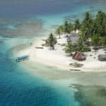 A tropical island with white sand beaches, turquoise shallow waters near shore, and dense palm trees; thatched huts line the beach and a small boat anchors at the shoreline.