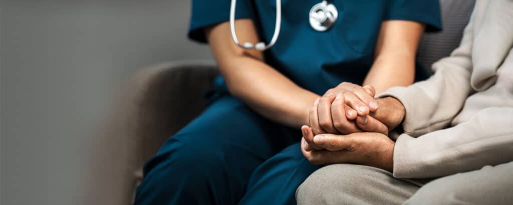 A healthcare professional in teal scrubs with a stethoscope sits holding the hands of a patient in a light-colored outfit, offering comfort.