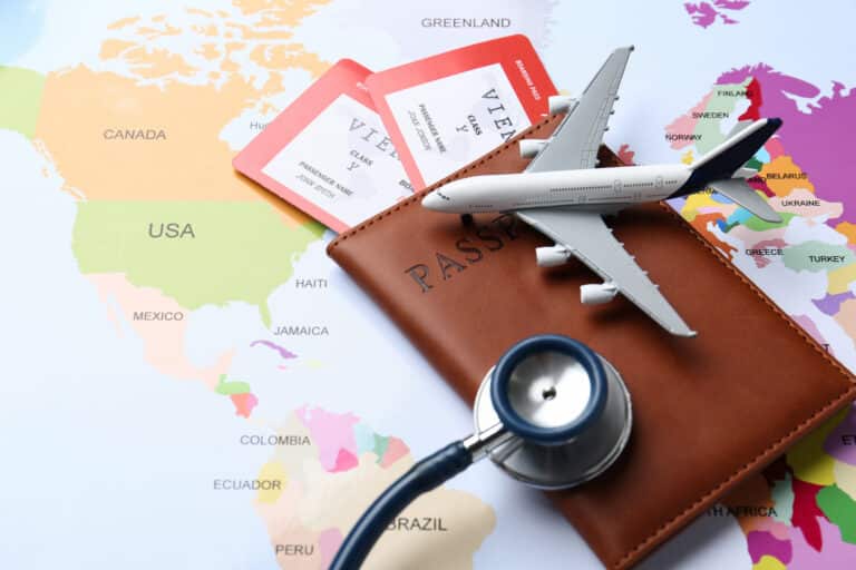 Airplane model, passport, and stethoscope resting on a brown wallet placed on a colorful world map with red boarding passes nearby.