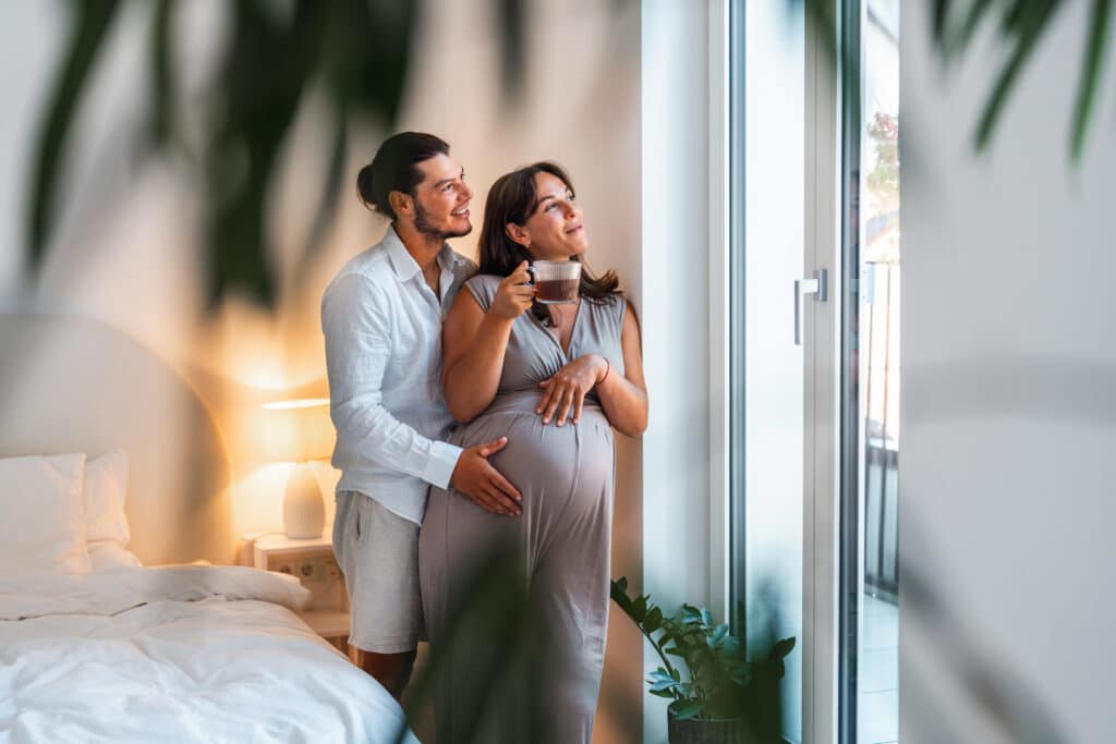 A pregnant woman in a gray dress stands by a window, holding a mug, while a man in a white shirt embraces her from behind in a softly lit bedroom.