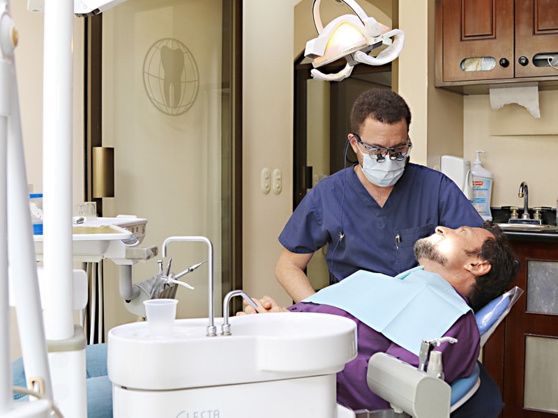 A dentist in blue scrubs and a mask leans over a patient lying in a dental chair, wearing a protective bib while a dental lamp hangs above.