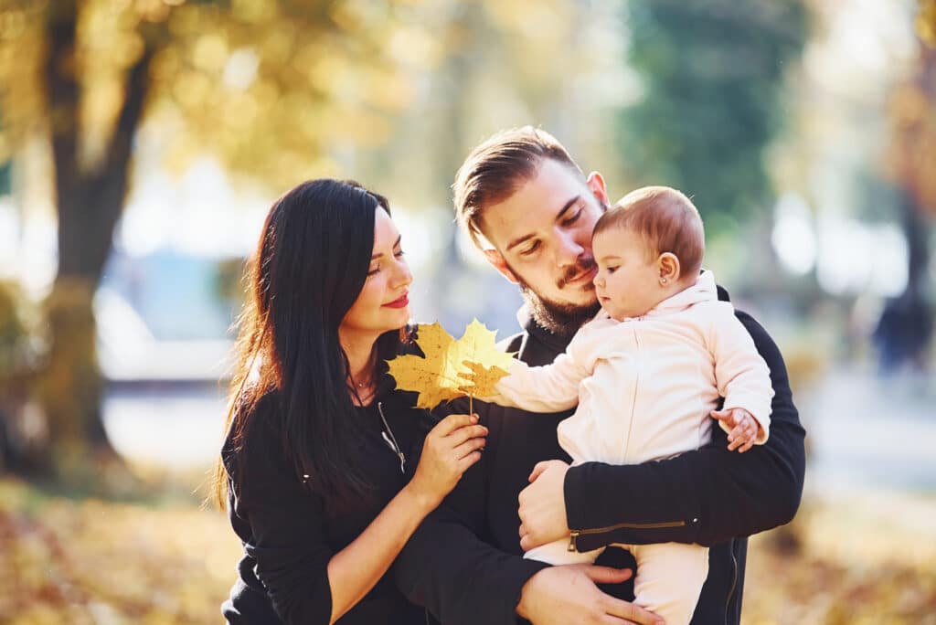 Family outdoors in autumn leaves; smiling parents hold a baby, woman with long dark hair and man with beard, warm sunlight.