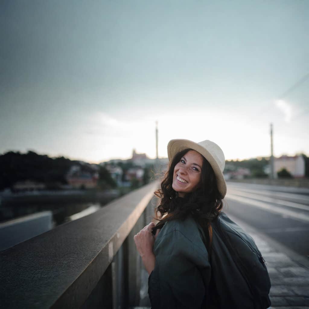 Smiling woman wearing a wide-brimmed hat near a railing on an outdoor walkway at sunset.