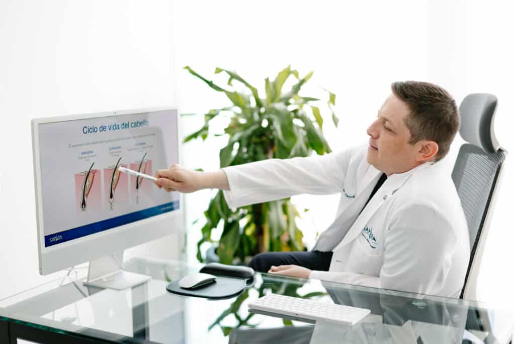 A man in a white coat sits at a glass desk, pointing a pen at a large computer monitor displaying a diagram titled “Ciclo de vida del cabello.”