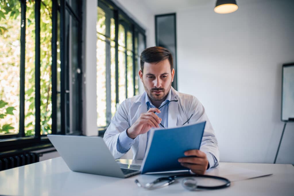 A man in a light blue shirt sits at a white table in a bright office, examining a document with a pair of glasses in hand; a laptop and stethoscope are on the table, with large windows and greenery outside.