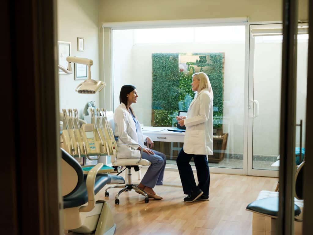 Two medical professionals in white coats chat in a bright clinic room with dental chairs, equipment, and a windowed door to an outdoor area.