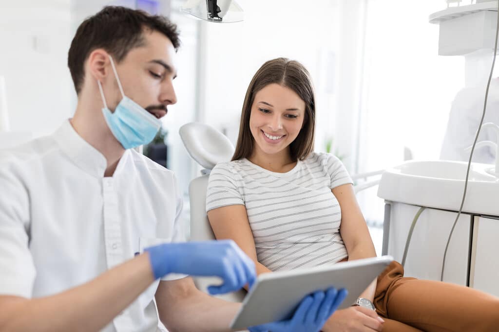 A male dentist wearing a face mask and blue gloves consults with a smiling female patient seated in a dental chair, holding a tablet.
