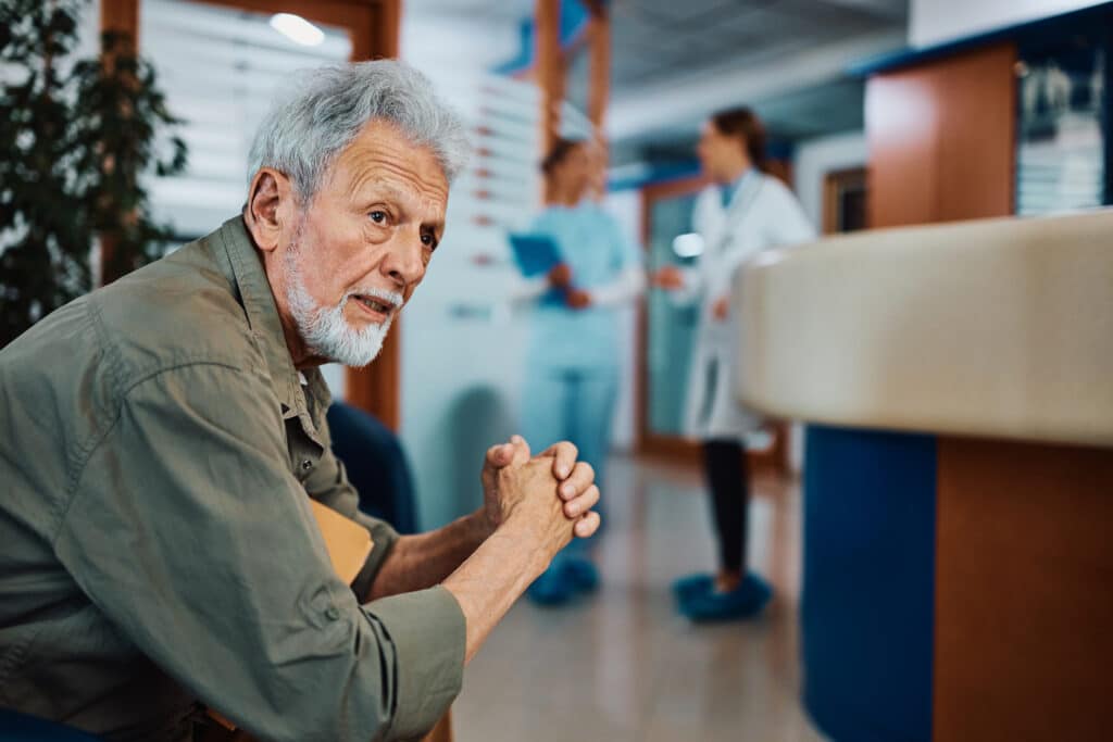 An elderly man with gray hair and a beard sits with hands clasped, looking pensively to the side in a bright, modern indoor setting.