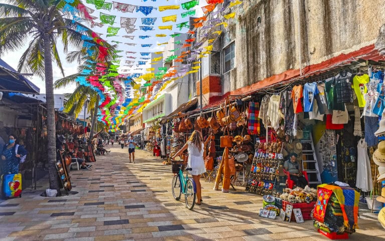 Colorful market street with stalls selling hats, bags, clothing and souvenirs; vibrant papel picado banners strung overhead; palm trees and sunny sky.