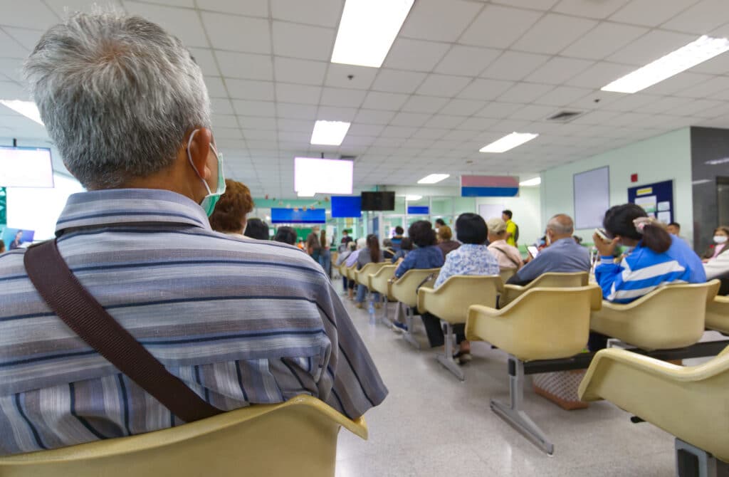 People seated in a waiting area or clinic with rows of beige chairs, many facing screens and a bright ceiling; foreground shows a man with gray hair and striped shirt from behind.