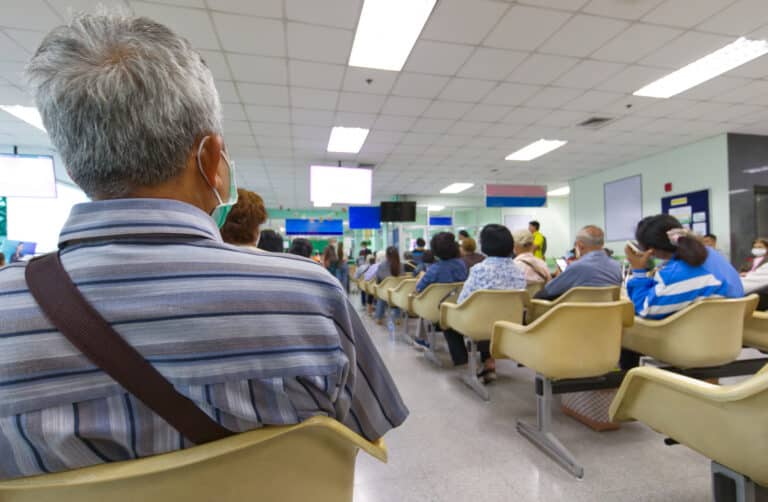 People seated in a waiting area or clinic with rows of beige chairs, many facing screens and a bright ceiling; foreground shows a man with gray hair and striped shirt from behind.