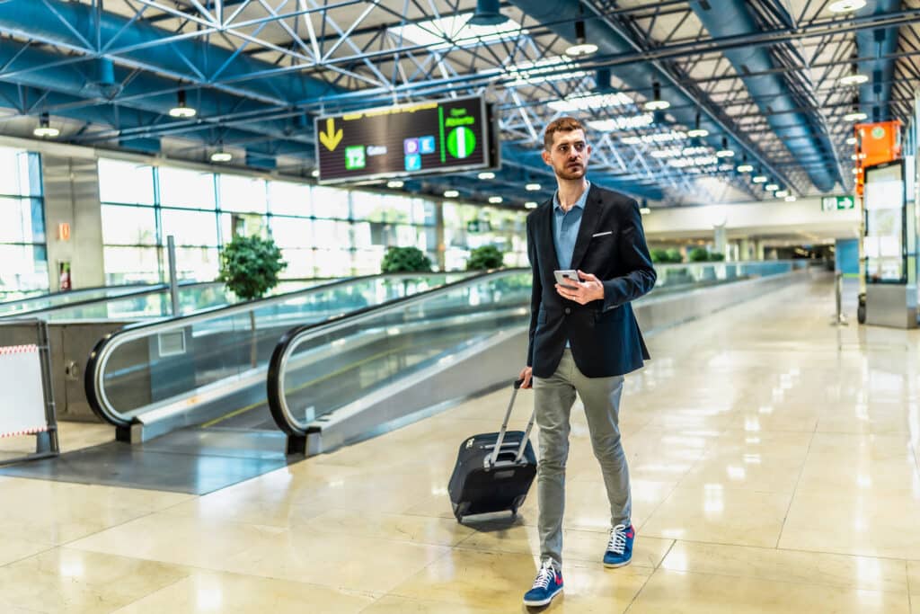 A man in a blazer and light pants walking in a modern airport, pulling a small rolling suitcase and looking to the side while holding a smartphone.
