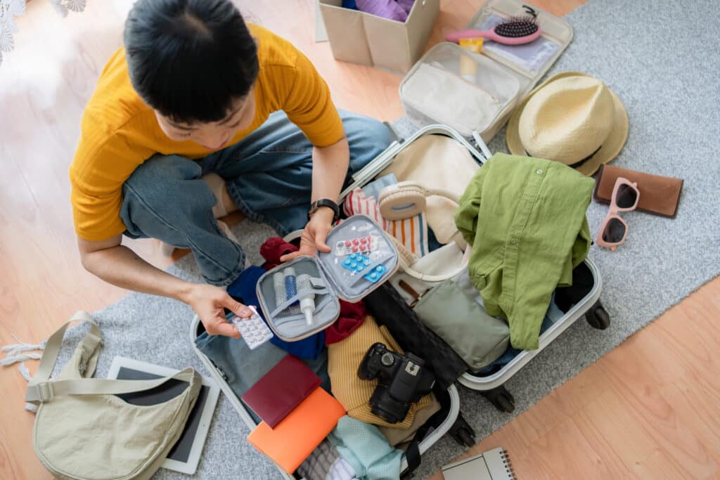 A person sorting through an open suitcase on a carpeted floor, surrounded by clothes, a hat, sunglasses, a camera, a notebook, and various travel items.