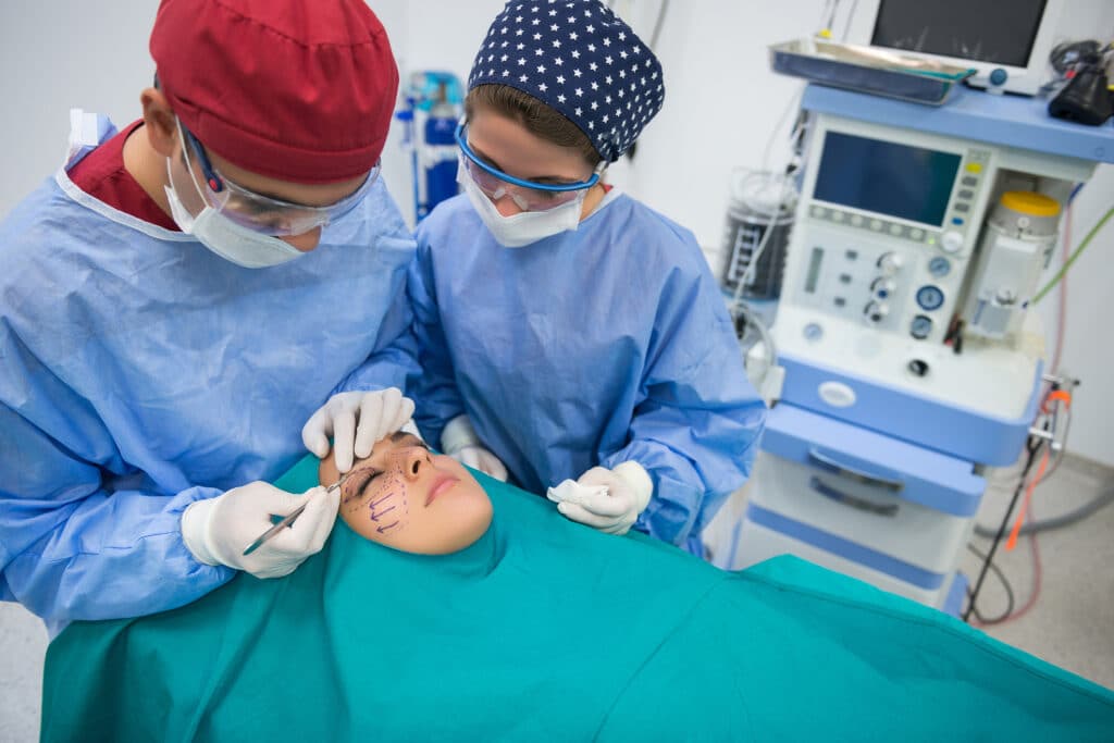 Two surgeons in protective gowns and masks operate on a patient, drawing marks on the face before a procedure in an operating room with medical equipment in the background.
