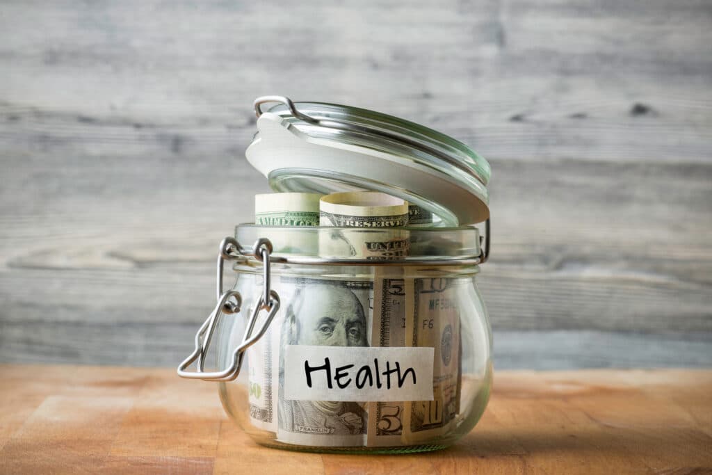 A glass mason jar filled with rolled dollar bills and labeled Health, sealed with a metal clasp and lid on a wooden surface.