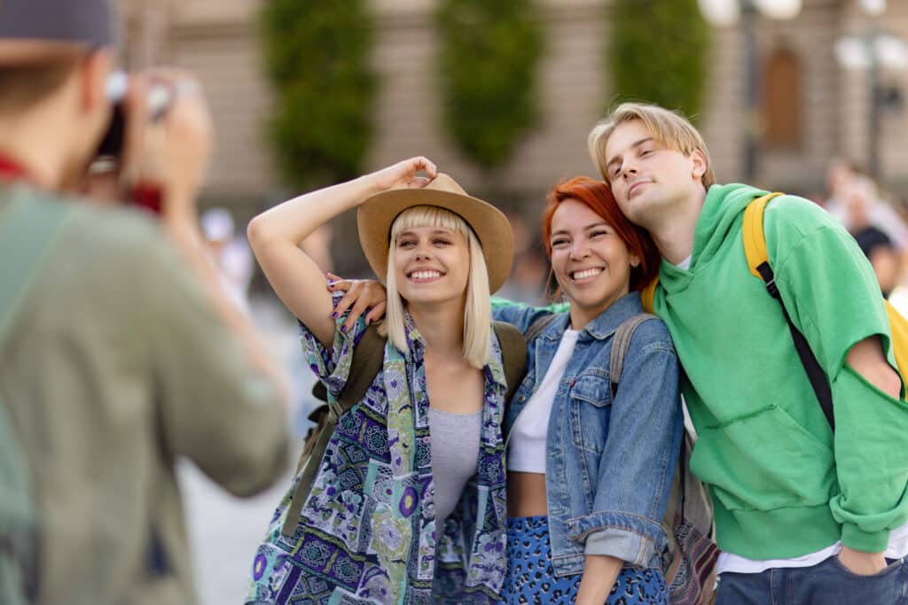 Three friends pose for a photo outdoors: a blond woman in a patterned, colorful cover-up and hat, a red-haired woman in a denim jacket, and a blond man in a bright green hoodie with a yellow backpack strap, smiling together.