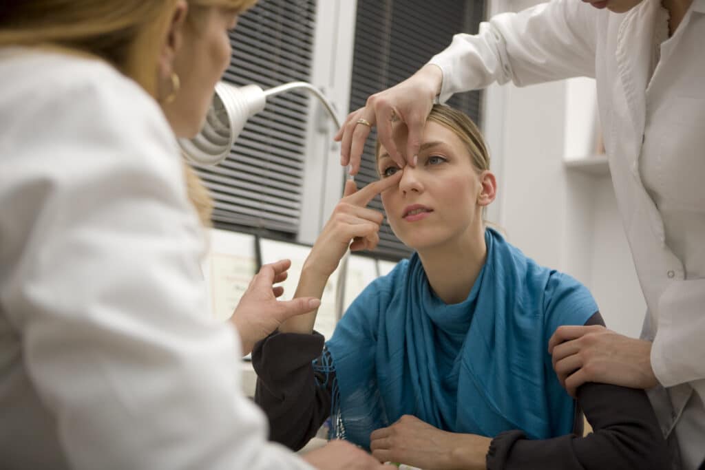 A young woman in a blue garment sits at a medical or clinical station while two clinicians in white coats adjust her eyelids, focusing on her eye.