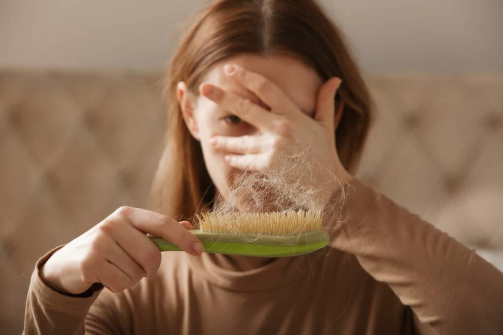 A person with light brown hair holds a green brush, covering part of their face with one hand while brushing hair.