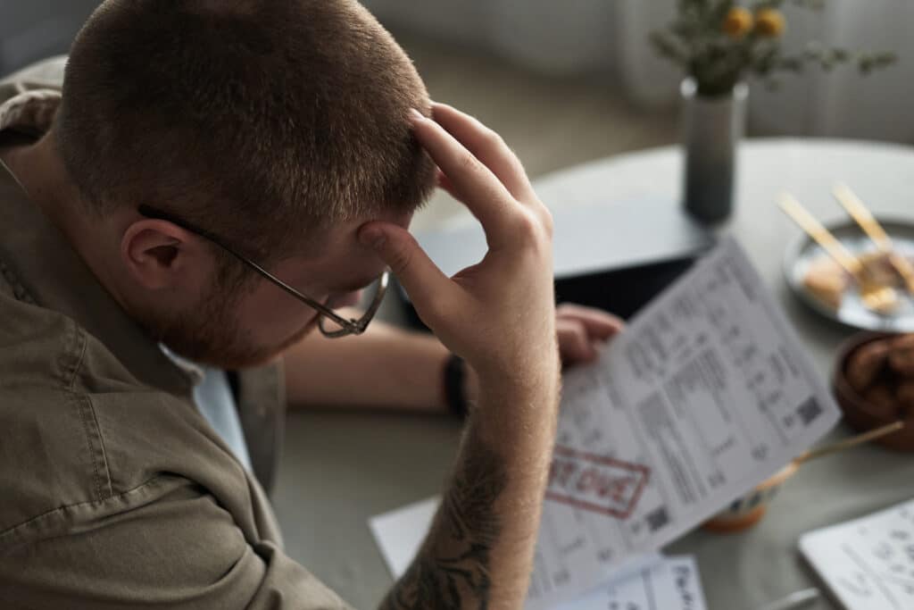A man with short hair and glasses rests his head in one hand while looking at papers on a table, with a plant and dish in the background.