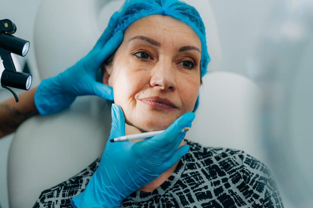 A close-up of a woman wearing a blue surgical cap and gloves, being pricked with a needle by a medical professional in blue gloves.